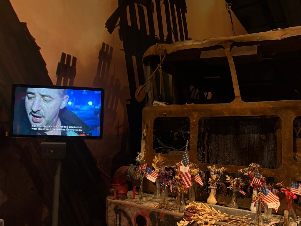 An exhibit featuring a damaged vehicle with flowers and small American flags arranged in front, alongside a screen displaying a man's testimony about an event.
