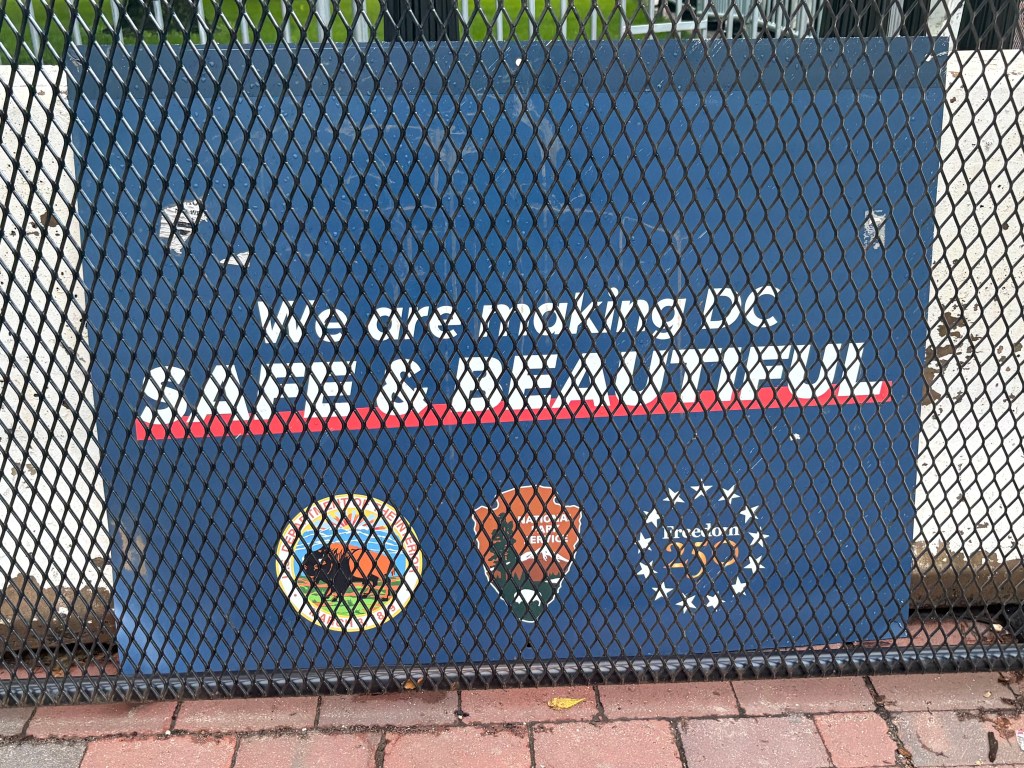 Sign reading 'We are making DC SAFE & BEAUTIFUL' with logos of city agencies, displayed behind a chain-link fence.