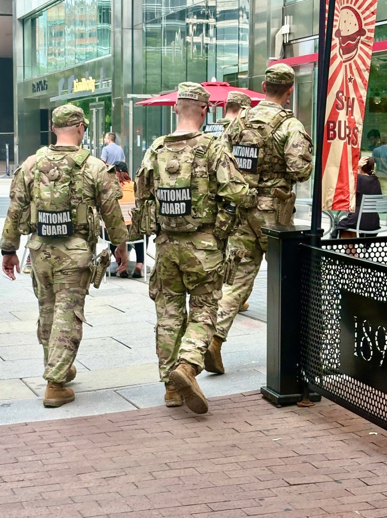 A group of four soldiers in camouflage uniforms with 'NATIONAL GUARD' patches walking in a city area, with pedestrians and outdoor seating visible in the background.