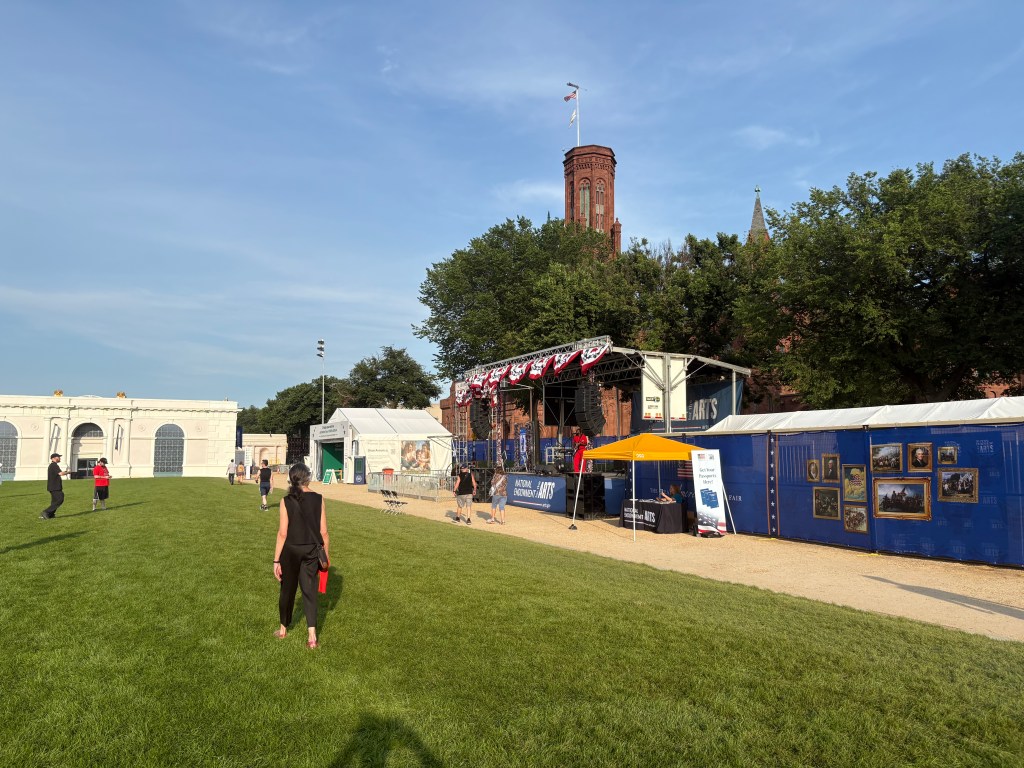 A grassy outdoor area with a stage decorated with bunting, surrounded by tents and people walking. In the background, a brick building with a clock tower under a clear blue sky.