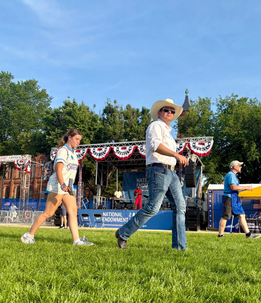 A man in a white shirt and cowboy hat walks on grass, while a girl in a baseball jersey passes by. A stage with patriotic decor is visible in the background.