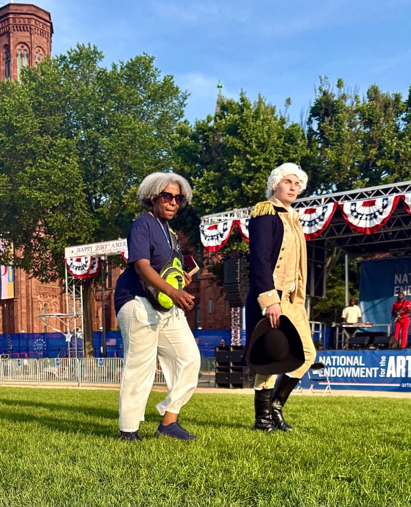A woman in sunglasses walks on grass, while a man dressed in a historical military uniform stands nearby. The background features a decorated stage for an event.