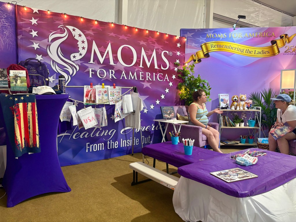 A trade show booth for Moms for America featuring a purple backdrop with the logo, a table with colorful craft supplies, and two women engaged in conversation.