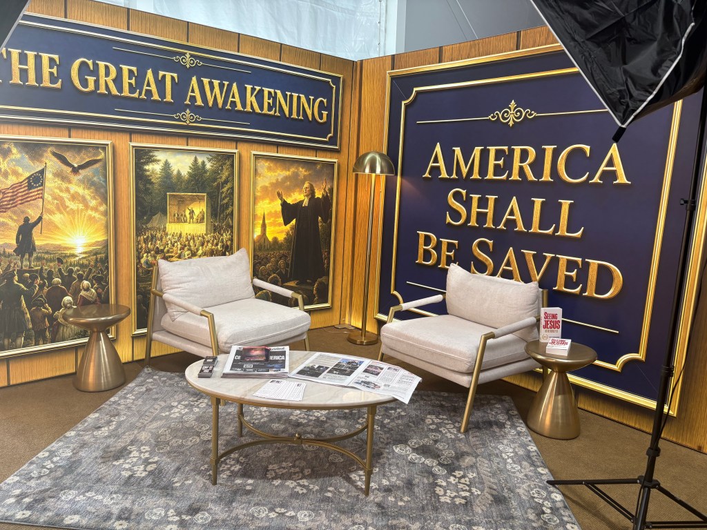 Interior setup featuring two modern chairs and a coffee table on a decorative rug, with a backdrop displaying the phrases 'THE GREAT AWAKENING' and 'AMERICA SHALL BE SAVED' alongside vibrant artwork depicting historical and patriotic themes.