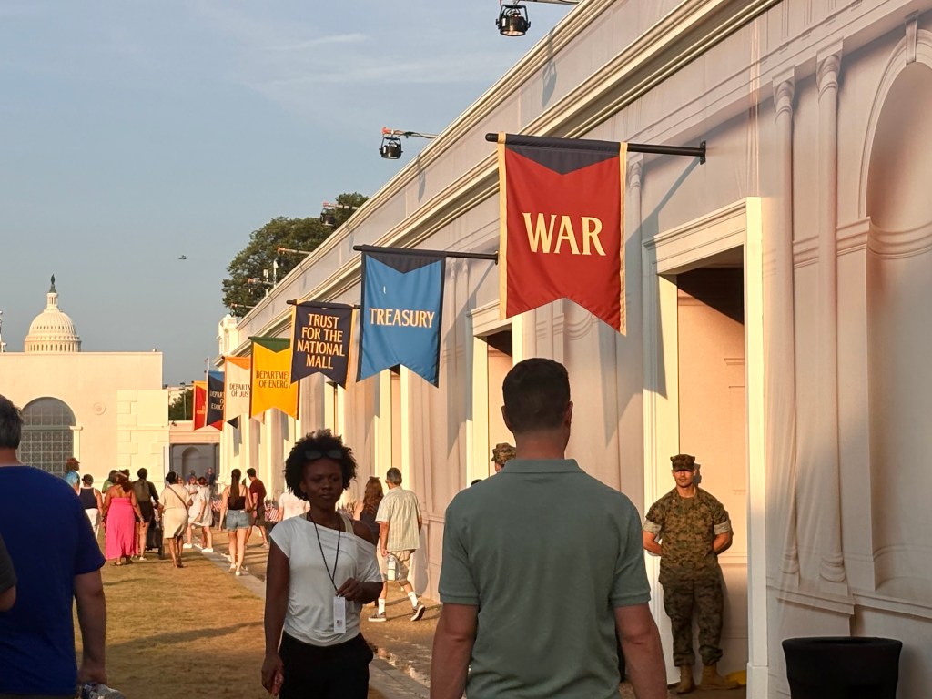 A diverse crowd walking past colorful banners labeled 'WAR', 'TREASURY', 'DEPARTMENT OF ENERGY', and 'TRUST FOR THE NATIONAL MALL' at an outdoor event, with the U.S. Capitol building visible in the background.