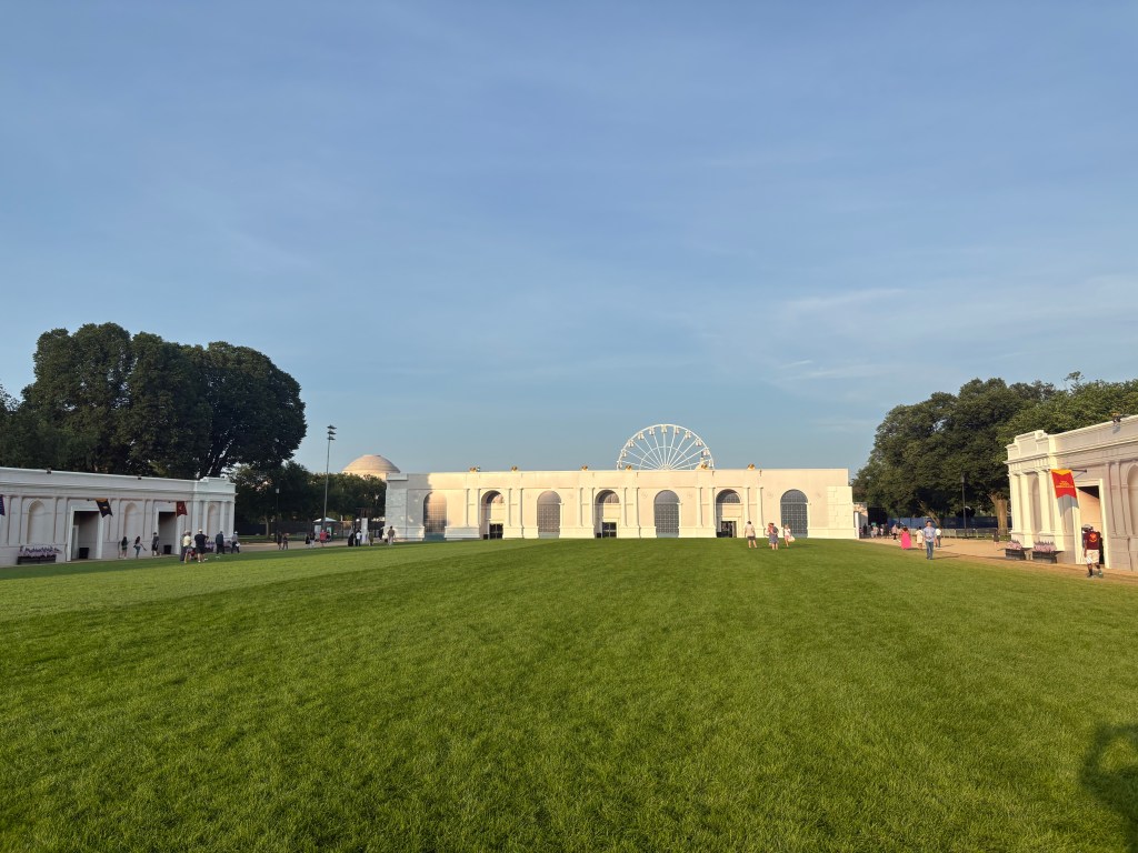 A wide view of an open grassy area with white architectural structures on either side, a ferris wheel in the background, and people enjoying the space under a clear blue sky.
