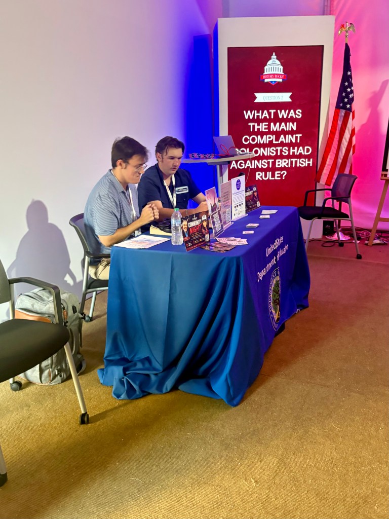 Two individuals seated at a blue table with informational materials, discussing a question about colonial complaints against British rule, backdrop features a large display with the same question.