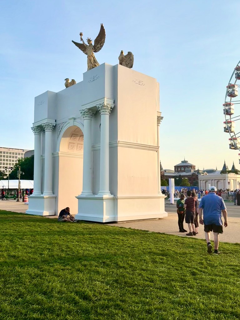 A large, white arch with golden statues of an eagle and a winged figure on top. People are walking nearby on a grassy area, with a Ferris wheel visible in the background.