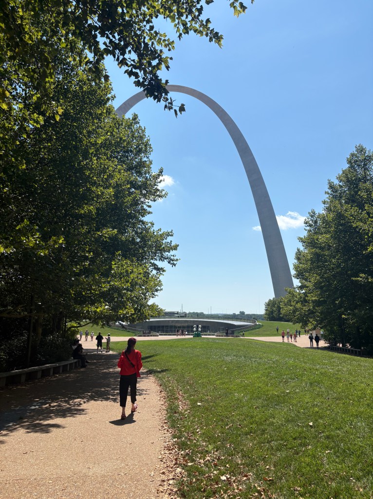 A person walking on a pathway towards the Gateway Arch, surrounded by trees and a clear blue sky.