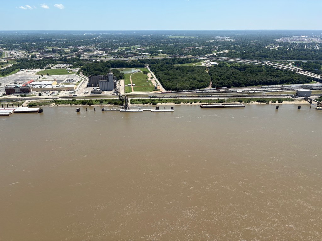Aerial view of a river with industrial buildings and greenery along the banks, showcasing urban landscape and transportation infrastructure.