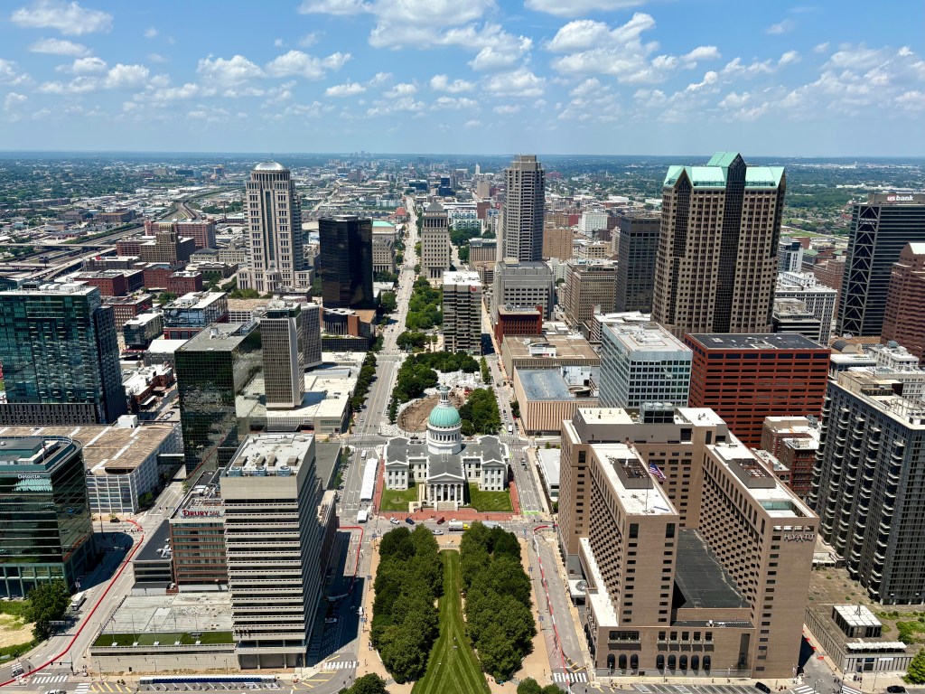 Aerial view of a city skyline featuring tall buildings, including modern glass skyscrapers and historic architecture, with clear blue skies and scattered clouds.