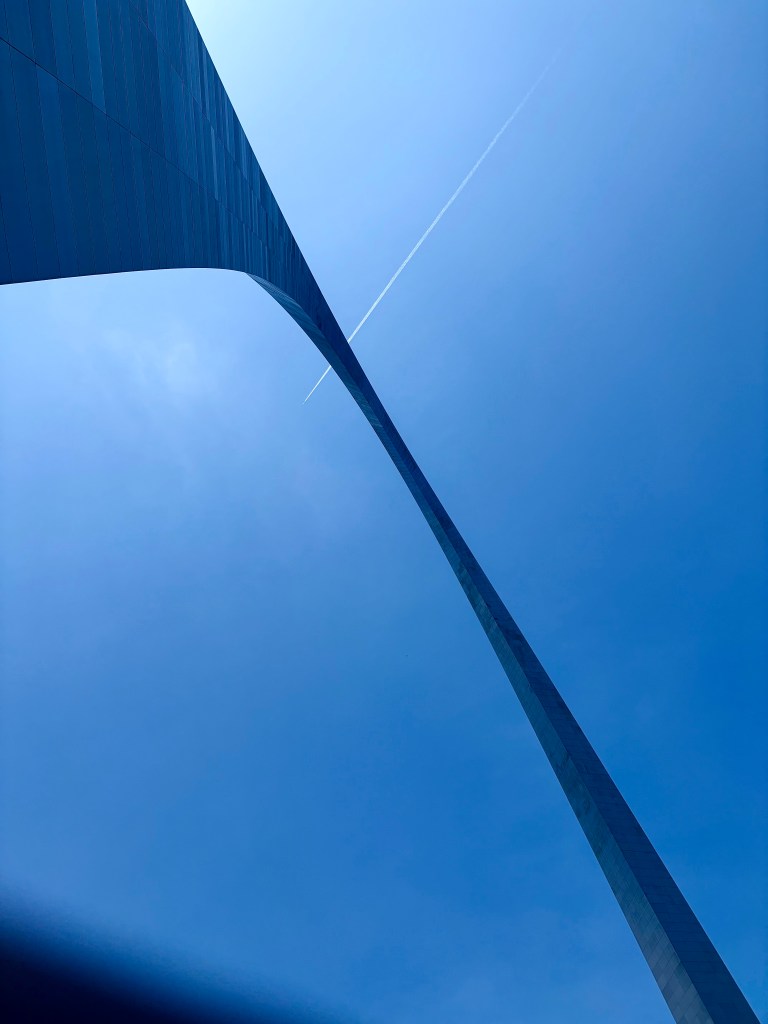 A towering monument with a sleek, curved design against a clear blue sky, featuring a faint airplane contrail above.
