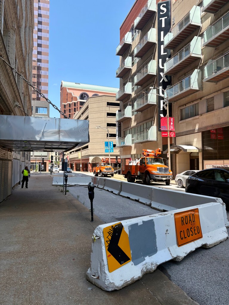 View of a city street with construction barriers blocking the road. In the background, there are high-rise buildings and orange construction vehicles parked. A pedestrian in a yellow vest is visible on the sidewalk.