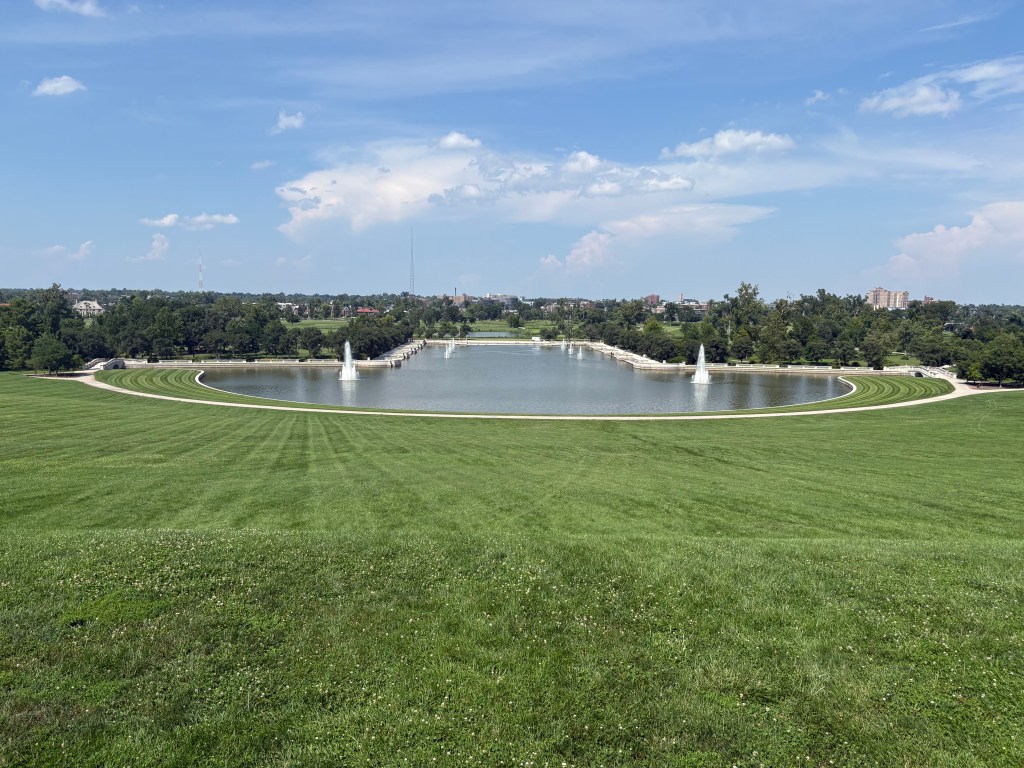 A panoramic view of a large, manicured park with a circular pond and fountains, surrounded by lush green grass and trees under a blue sky.