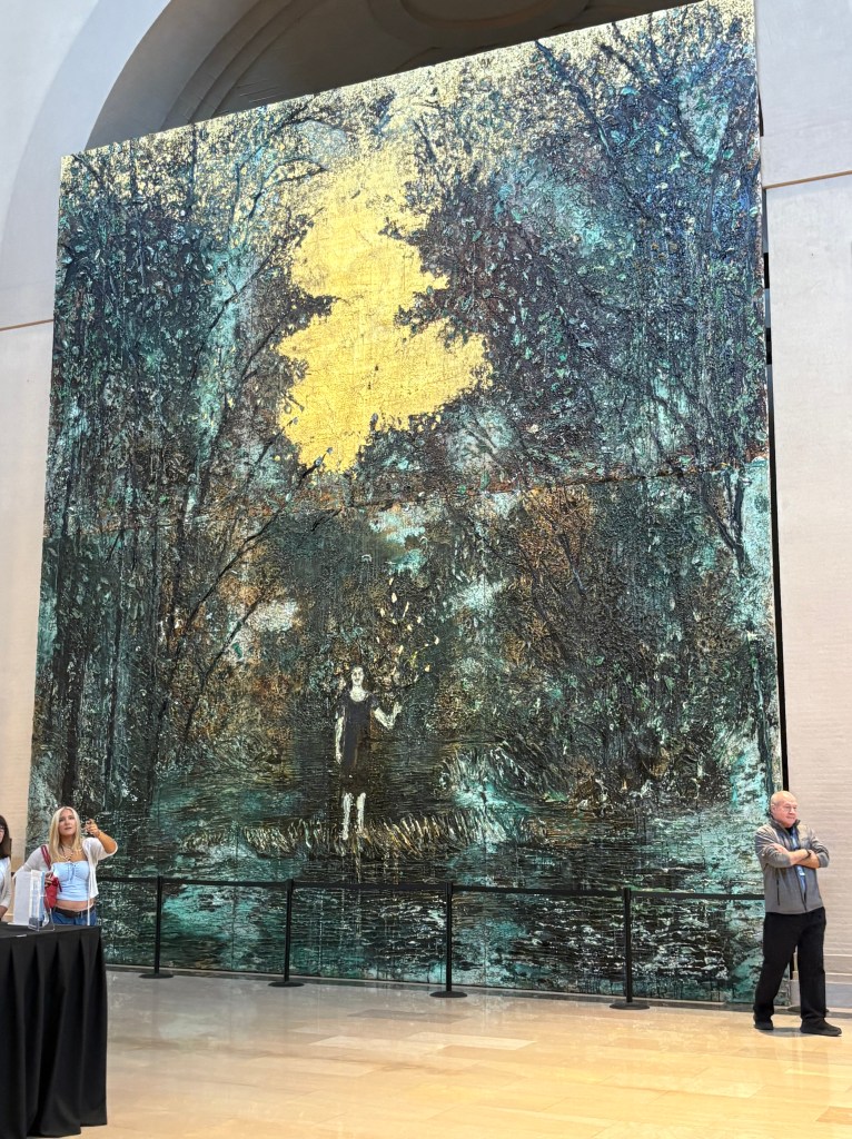 A large mural depicting a serene landscape with muted colors, featuring trees and a figure standing amidst the scenery, highlighted by a bright yellow area. Two visitors are standing in front of the artwork in an indoor gallery space.