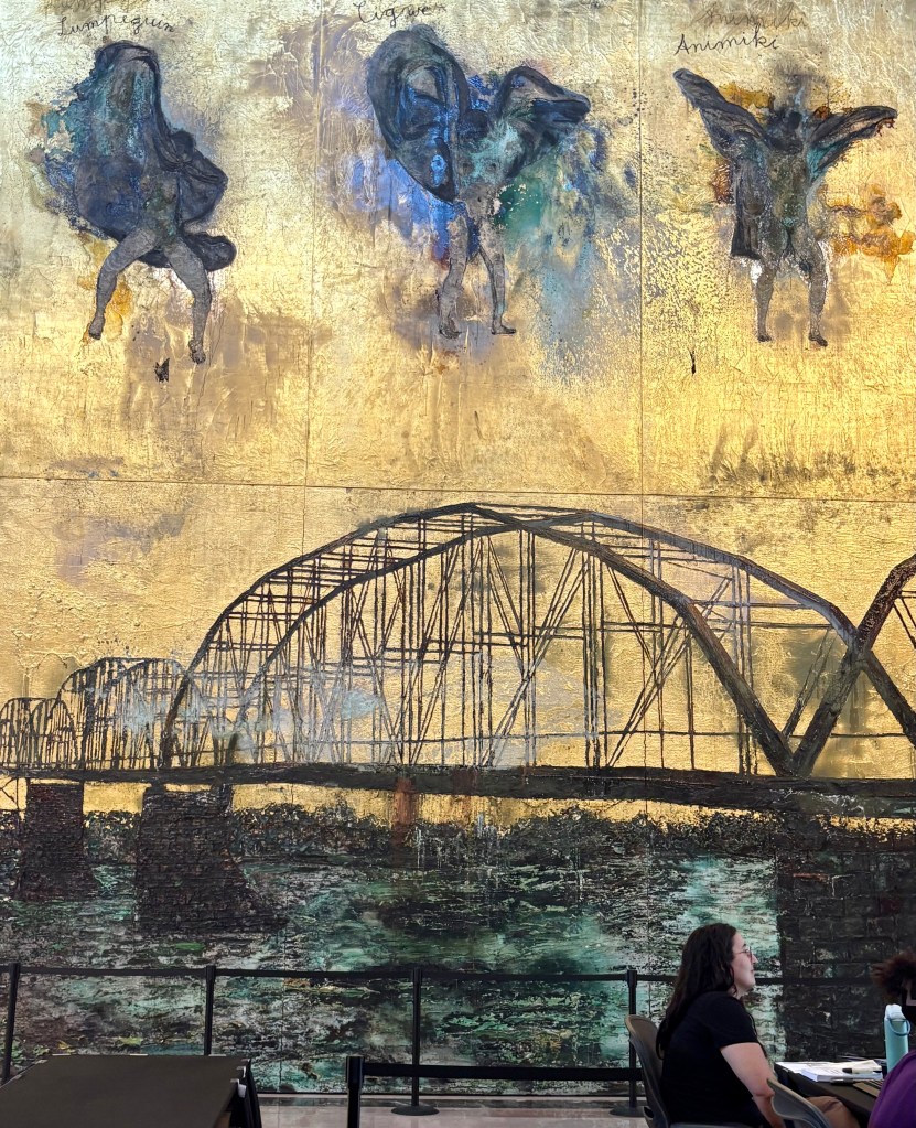 A large mural featuring abstract figures with wings against a gold background, depicting a bridge over water with a person sitting at a table in the foreground.