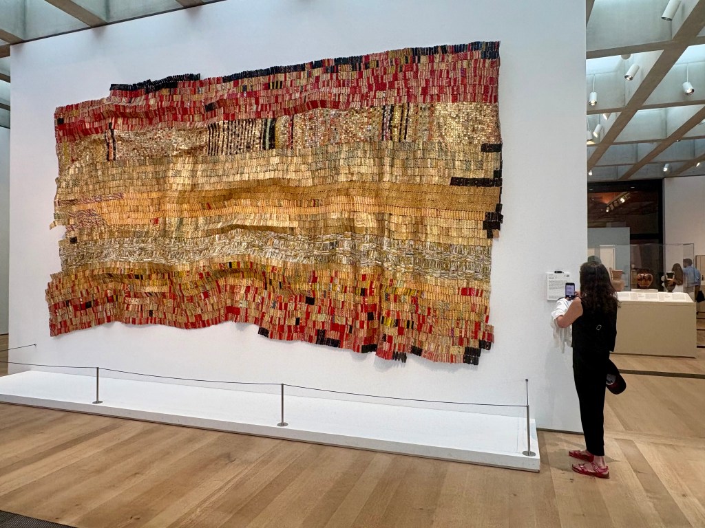 An art installation featuring a large tapestry made of various colored strips, predominantly gold, red, and black, displayed in a gallery. A person is observing the artwork, standing beside it.