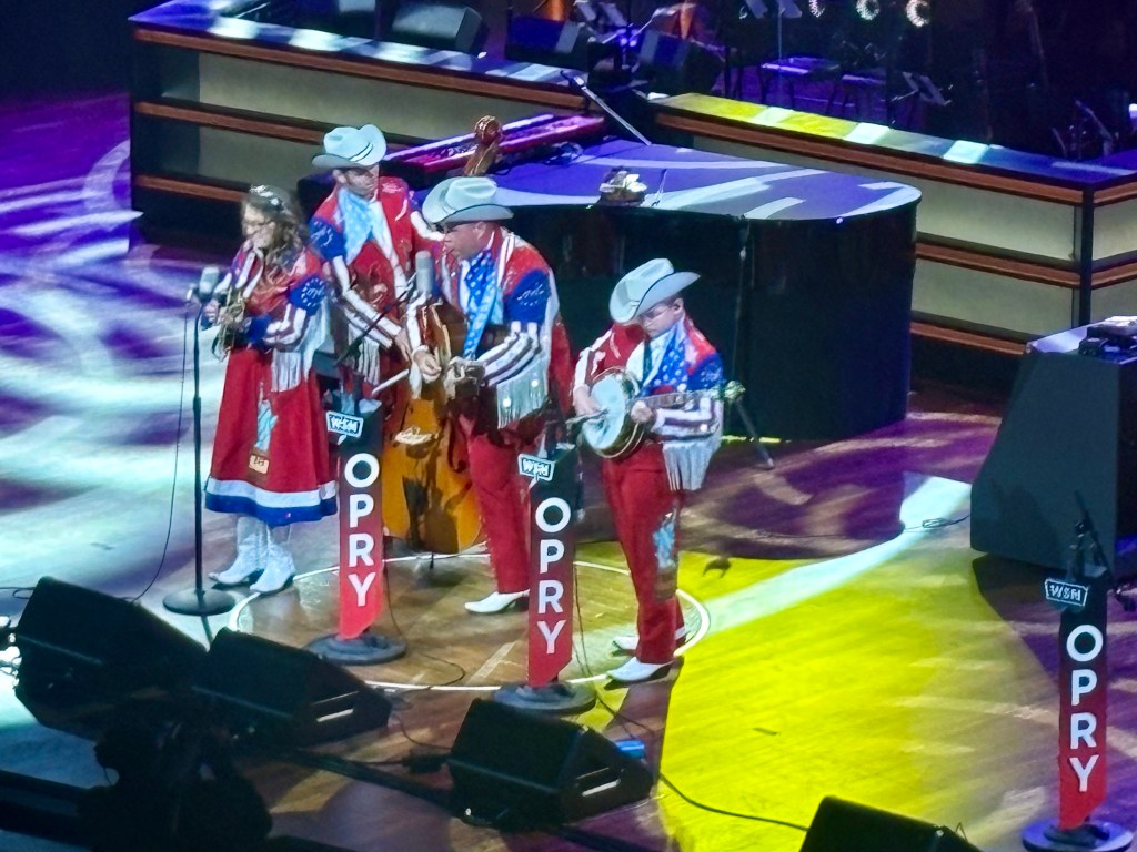 A bluegrass band performing on stage wearing colorful outfits, with instruments including a banjo and a double bass, at the Grand Ole Opry.