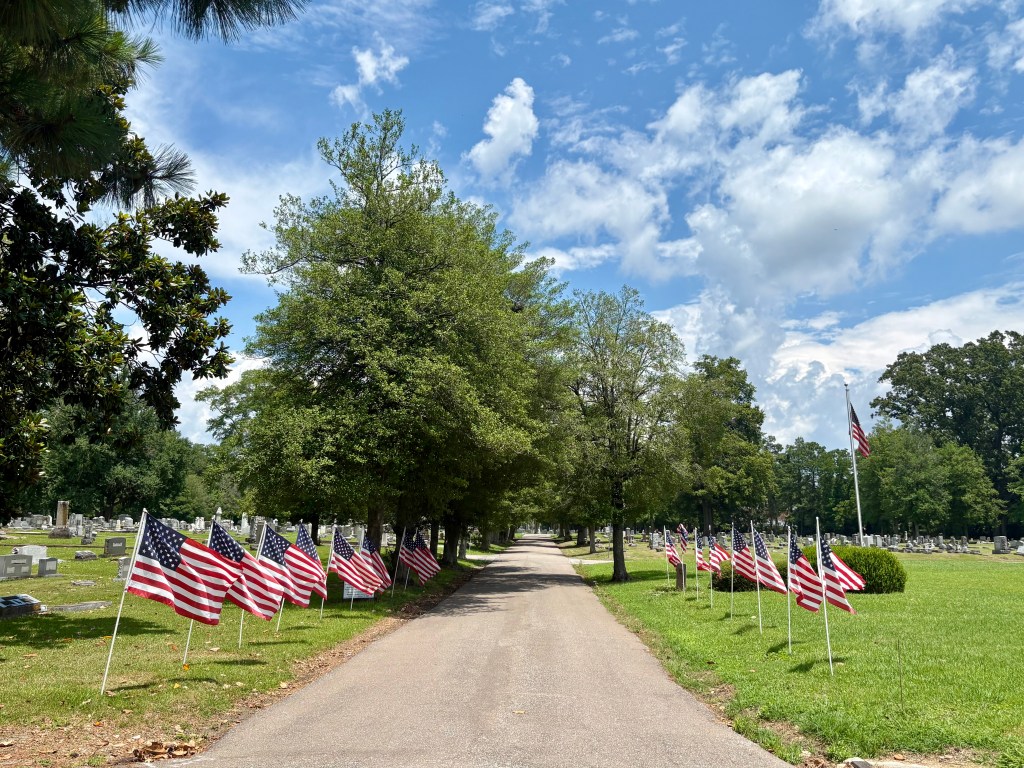 A peaceful cemetery scene featuring a pathway flanked by American flags and trees under a blue sky with scattered clouds.