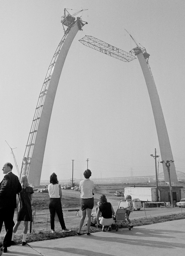 Viewers watching the construction of a large arch structure, with scaffolding and cranes visible, in black and white.