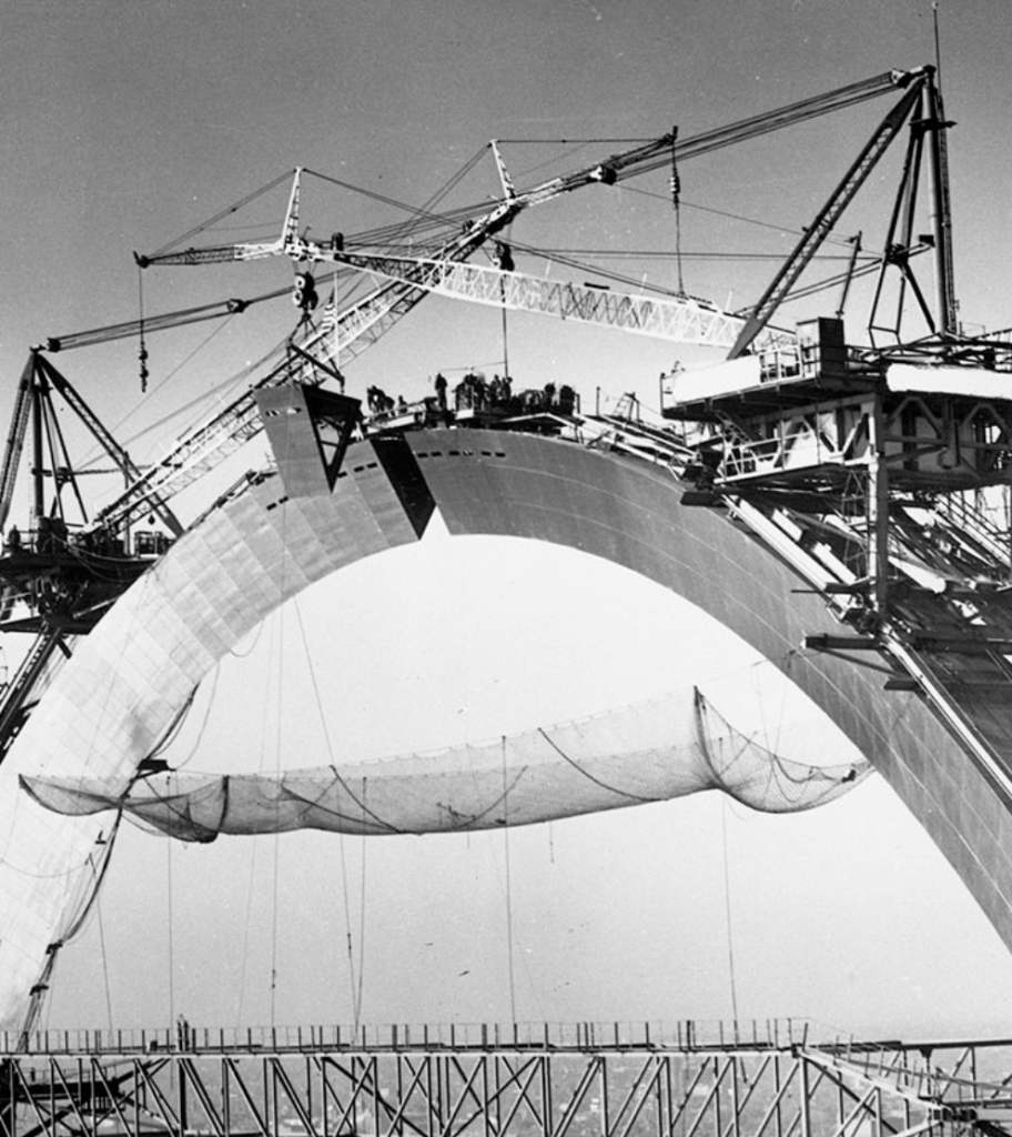 Black and white image of a large arch bridge under construction, featuring cranes and workers on top, with a safety net hanging underneath the arch.