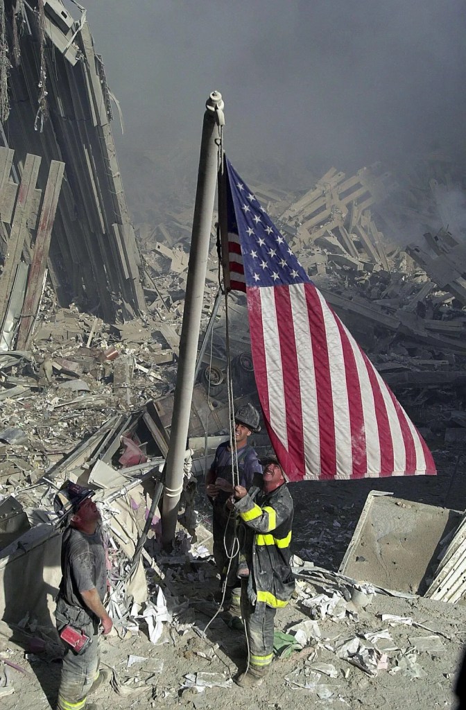 Firefighters raising an American flag at the site of destruction, surrounded by debris and smoke.